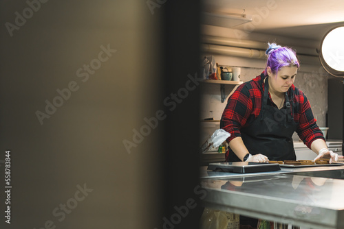 Female baker wearing red plaid shirt and black apron is preparing desserts on a metal counter in a kitchen. Blurred foreground . High quality photo © PoppyPix