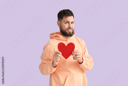 Young bearded man with paper heart for Valentine's Day on lilac background