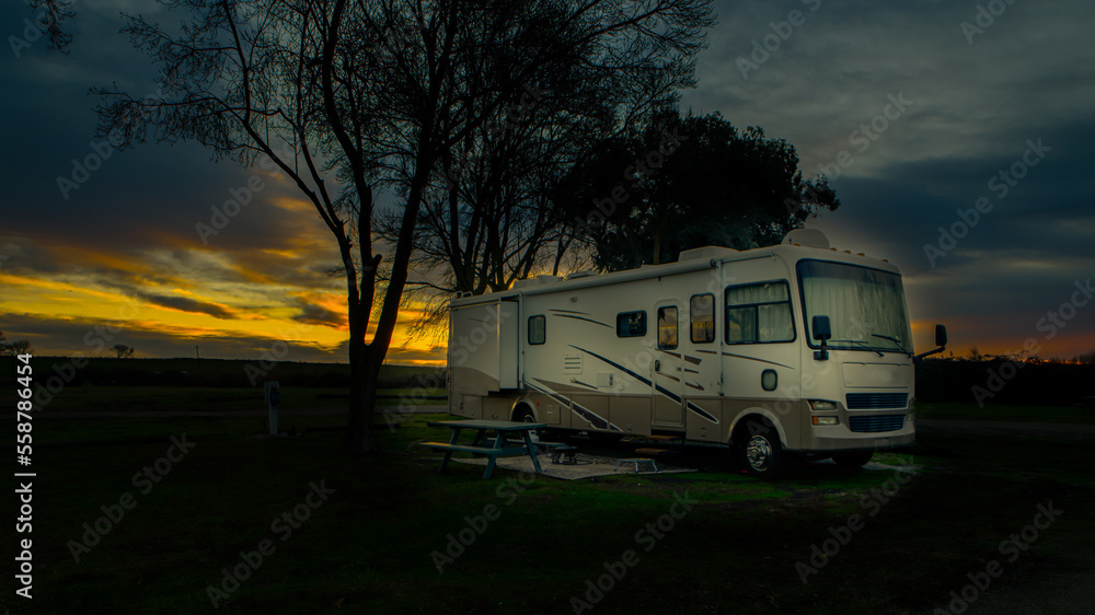Rv camper parked under cloudy sunrise sky and trees at campsite Rv camper parked under cloudy sunrise sky and trees at campsite