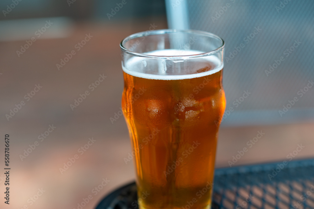 A clear pint drinking glass filled with cold froth from a lager ale. The Irish red ale pint sits on the edge of a metal patio table at a microbrewery. There are tables and chairs in the background. A clear pint drinking glass filled with cold froth from a lager ale. The Irish red ale pint sits on the edge of a metal patio table at a microbrewery. There are tables and chairs in the background.