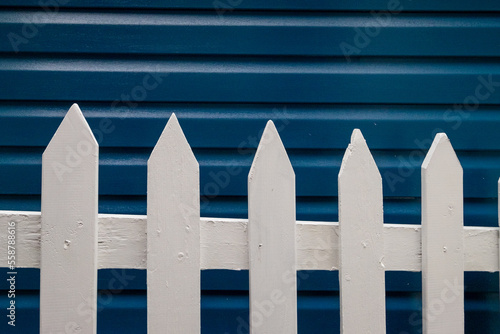 A narrow navy blue wood Cape Cod siding exterior of a vintage building with white trim around the window. There's a country style white wooden picket fence in the foreground with lats and rails.