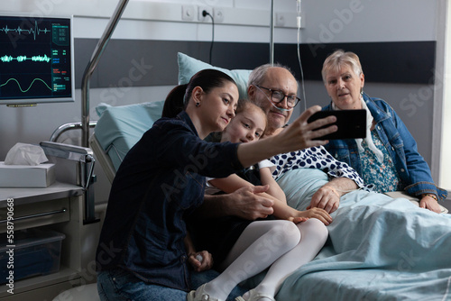 Sick citizen adult daughter taking cell phone self portrait with family members in hospital monitoring room. Old bedridden male patient family shooting selfie in sanatorium room.