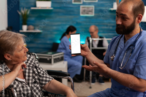 Male nurse showing white cellular screen while assisting elderly woman in nursing home. Professional services dedicated to the care of retired persons with special needs.