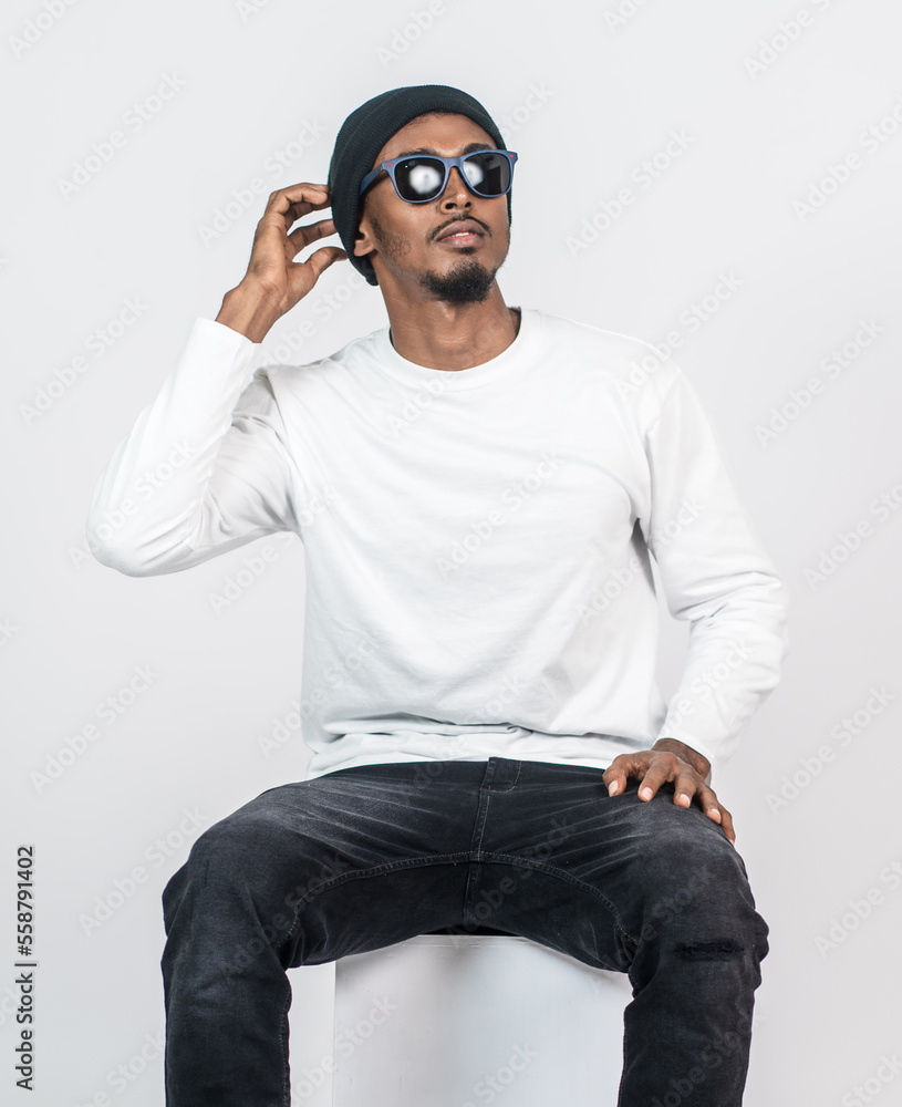 A young man wearing white blank shirt, with a hat and glasses sitting down to a chair A young man wearing white blank shirt, with a hat and glasses sitting down to a chair