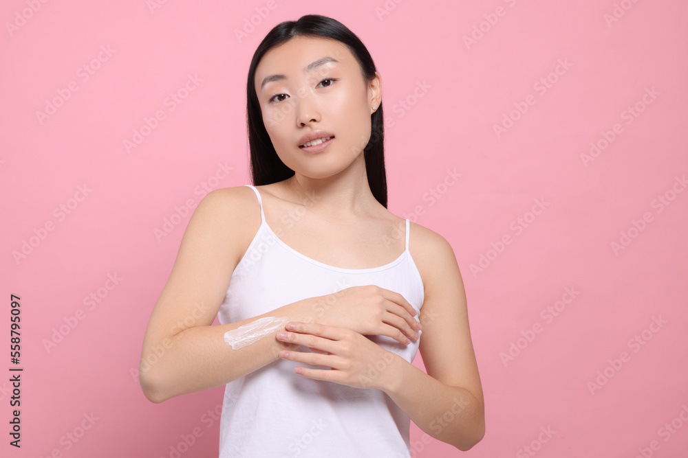 Beautiful young Asian woman applying body cream onto arm on pink background Beautiful young Asian woman applying body cream onto arm on pink background