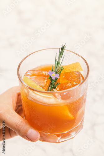 An orange color cocktail in a rocks glass decorated with orange peel, rosemary.
