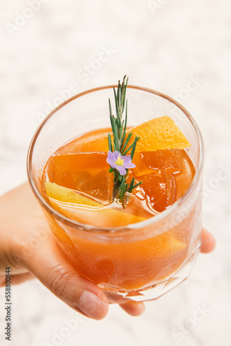 A hand holding orange color cocktail in a rocks glass decorated with orange peel, rosemary and purple flower.