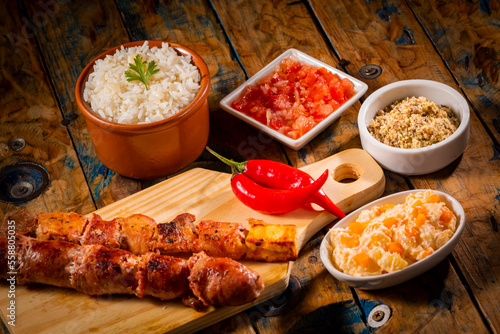 Espetinhos de churrasco em tábua de madeira com acompanhamentos, arroz, vinagrete, farofa e maionese