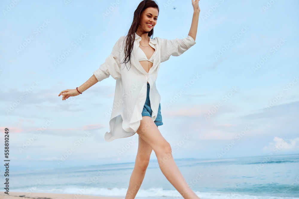 Happy tanned woman in white swimsuit shirt and denim shorts walks on the beach on the sand by the ocean with wet hair after swimming, sunset light and pink clouds in Bali Happy tanned woman in white swimsuit shirt and denim shorts walks on the beach on the sand by the ocean with wet hair after swimming, sunset light and pink clouds in Bali