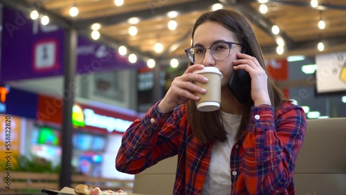 A young woman in a cafe is talking on the phone and drinking a hot drink. Girl in a shirt and glasses in a cozy cafe.