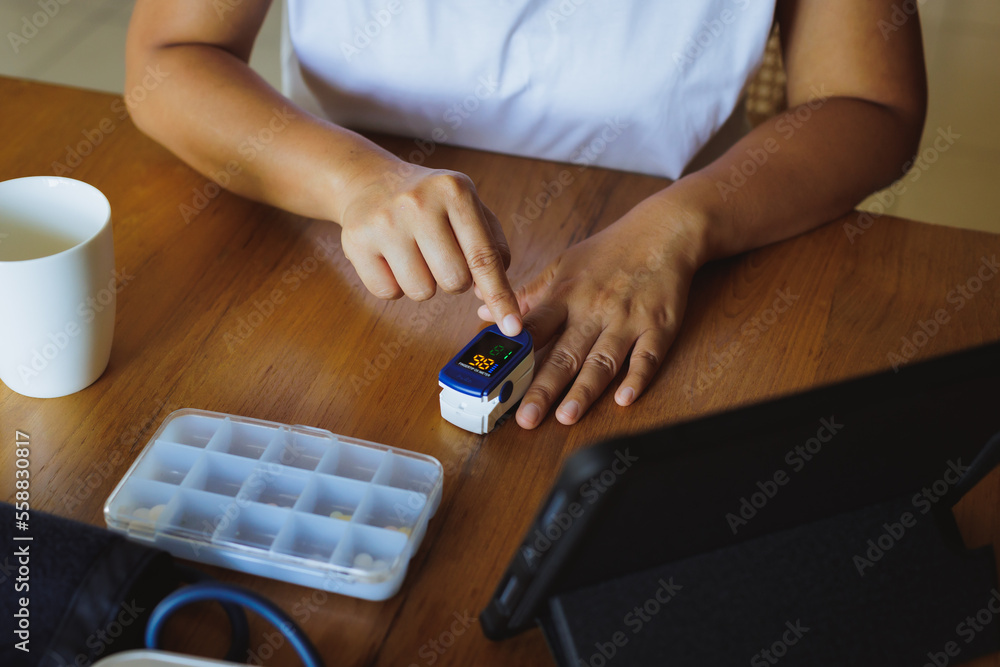 Asian woman self testing for blood oxygen levels with pulse oximeter monitor on her finger at home and record result in the tablet. Asian woman self testing for blood oxygen levels with pulse oximeter monitor on her finger at home and record result in the tablet.