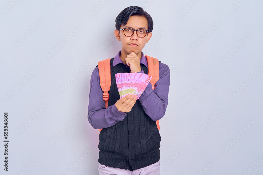 Pensive young Asian student man in casual clothes with backpack Holding fan of cash money in rupiah banknotes isolated on gray background. High school university college concept Pensive young Asian student man in casual clothes with backpack Holding fan of cash money in rupiah banknotes isolated on gray background. High school university college concept