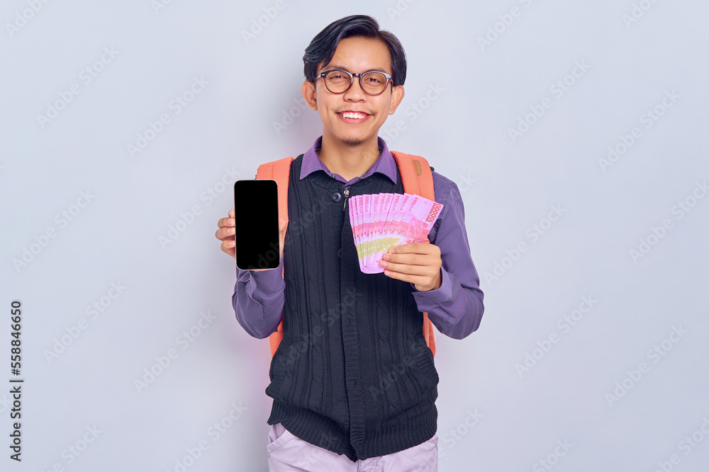 Smiling young Asian student man in casual clothes with backpack holding mobile phone with blank screen and cash money in rupiah banknotes isolated on gray background Smiling young Asian student man in casual clothes with backpack holding mobile phone with blank screen and cash money in rupiah banknotes isolated on gray background