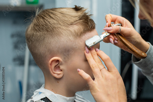 A boy is sitting in a barbershop, doing his hair with a razor for a haircut