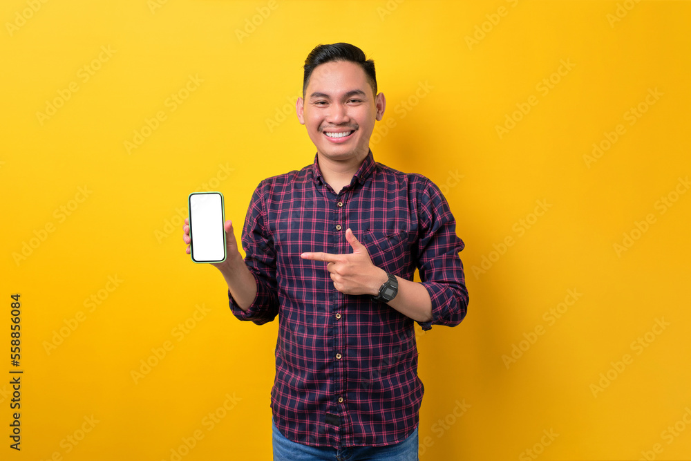 Smiling young Asian man pointing index finger at empty smartphone screen isolated on yellow background. Advertising new mobile app, mockup concept Smiling young Asian man pointing index finger at empty smartphone screen isolated on yellow background. Advertising new mobile app, mockup concept