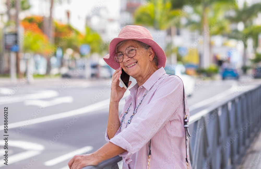 Cheerful attractive senior woman with summer hat standing in the city street using mobile phone