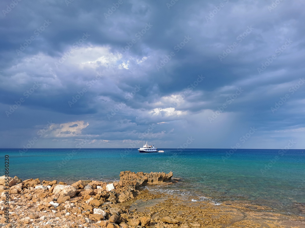 The stone coast of the Mediterranean Sea against the backdrop of the sea with a walking white ship and a dramatic sky. The stone coast of the Mediterranean Sea against the backdrop of the sea with a walking white ship and a dramatic sky.