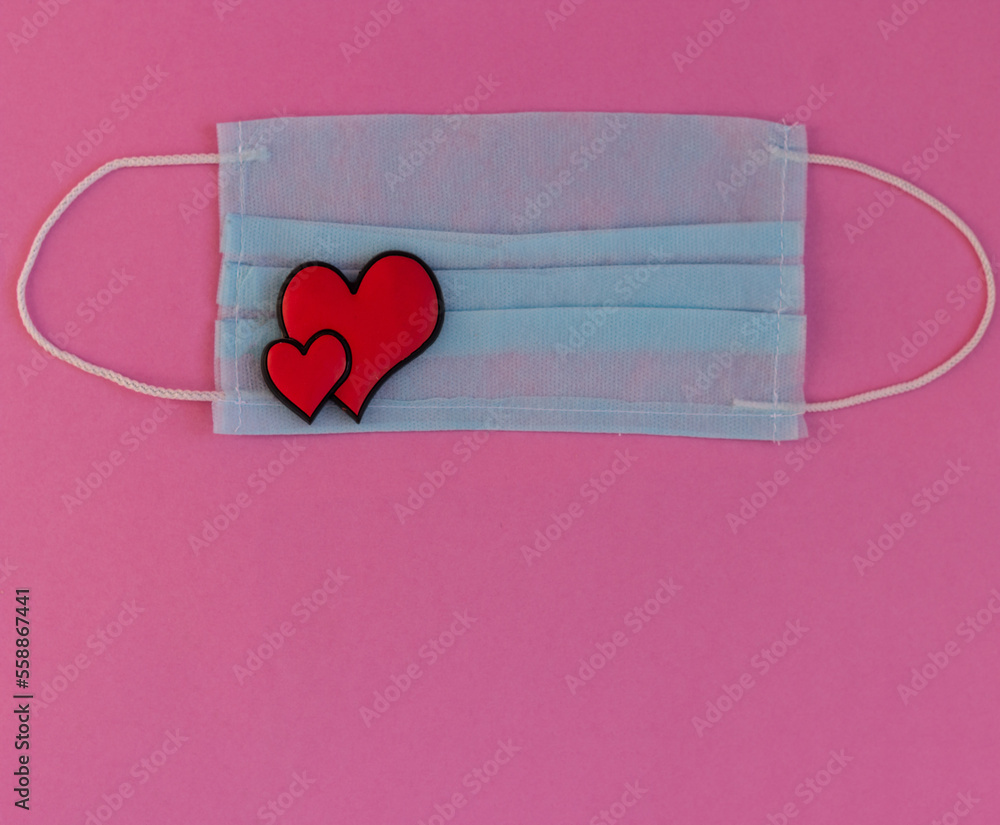 Two bright red hearts lie in the lower left corner on a medical mask on a pink background. The concept of Valentine's Day during a pandemic. With copy space. Close-up.