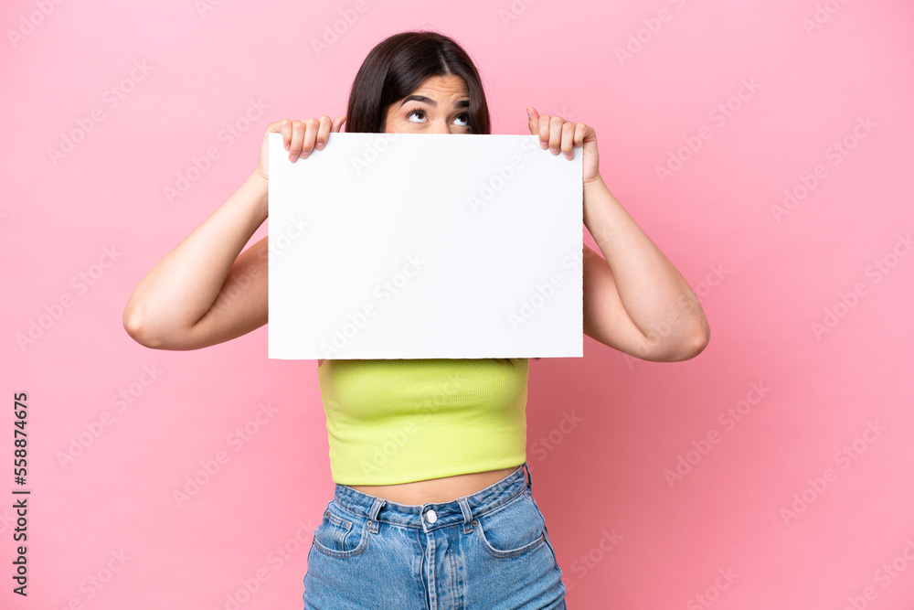 Young Brazilian woman isolated on pink background holding an empty placard and hiding behind it Young Brazilian woman isolated on pink background holding an empty placard and hiding behind it