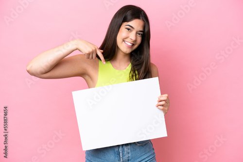 Young Brazilian woman isolated on pink background holding an empty placard with happy expression and pointing it