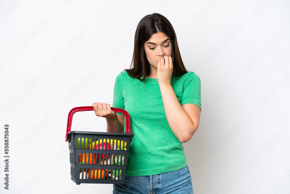 Young Brazilian woman holding a shopping basket full of food isolated on white background having doubts Young Brazilian woman holding a shopping basket full of food isolated on white background having doubts