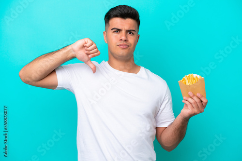 Young caucasian man catching french fries isolated on blue background showing thumb down with negative expression © luismolinero