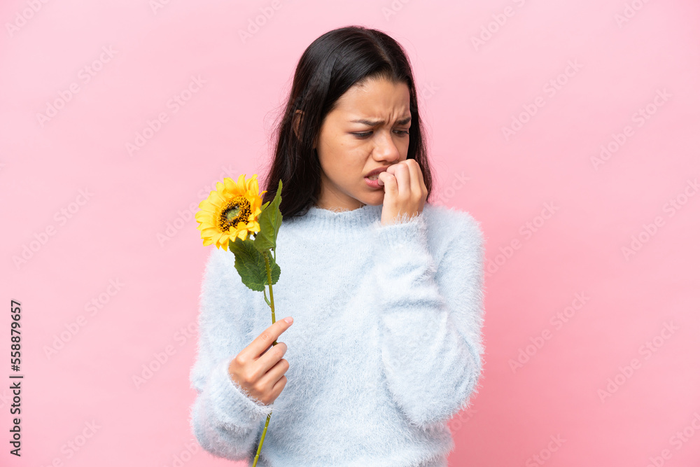 Young Colombian woman holding sunflower isolated on pink background having doubts Young Colombian woman holding sunflower isolated on pink background having doubts