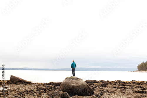 A young woman in a teal jacket and jeans ooks across Desolation Sound from a large boulder at low tide on a gray day. photo