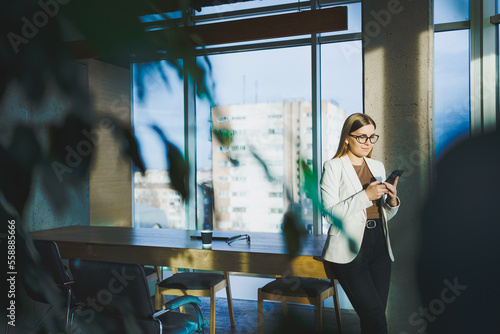 A successful young woman in glasses and a jacket is standing in the office and happily talking on the phone. A young manager works in the office and manages the business remotely
