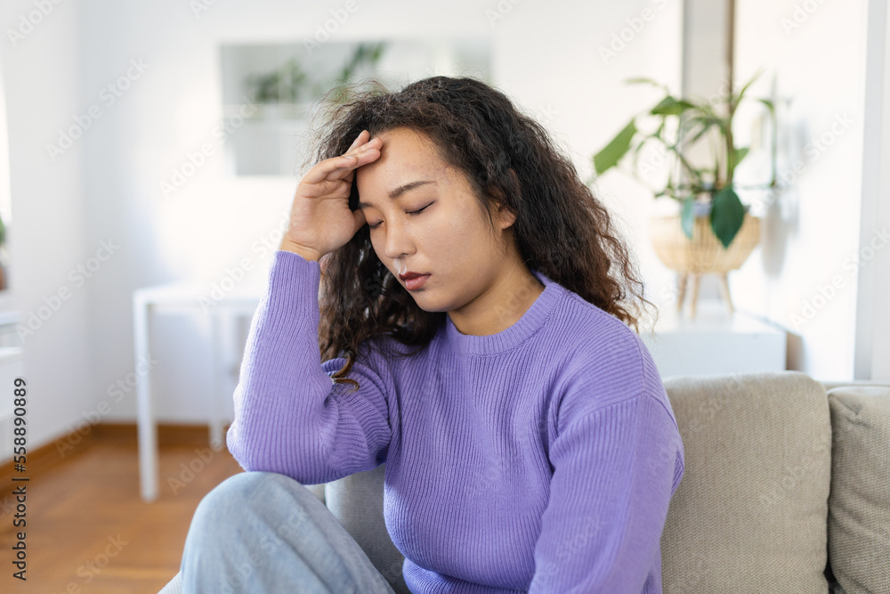 Portrait of a young Asian woman sitting on the couch at home with a headache and pain. Beautiful woman suffering from chronic daily headaches. Sad woman holding her head because sinus pain Portrait of a young Asian woman sitting on the couch at home with a headache and pain. Beautiful woman suffering from chronic daily headaches. Sad woman holding her head because sinus pain