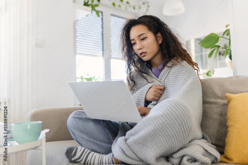 Asian woman wrapped in a cozy blanket sitting on the sofa and working on her laptop Asian woman wrapped in a cozy blanket sitting on the sofa and working on her laptop