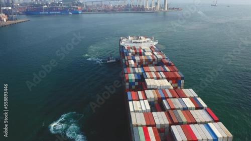 Giant container vessel pushed towards the dock by tugboats for unloading, with the Stonecutters bridge and container port in the background in Hong Kong. Approaching tilt-down drone shot