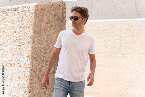 Handsome hipster guy wearing white empty t-shirt and sunglasses is standing in a building site. Vertical mock-up.