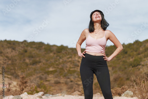 young latin woman posing standing smiling with eyes closed and hands on hips, in an arid valley.