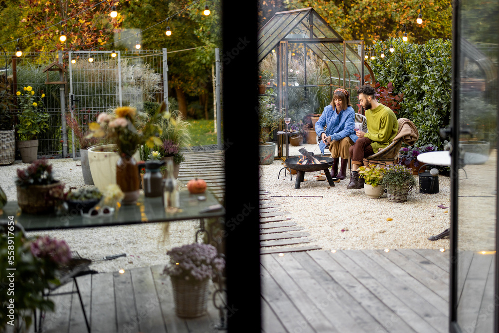 Young stylish couple grilling food and warming up while sitting together by the fire, spending autumn evening time at cozy atmosphere in garden. Wide view through the window