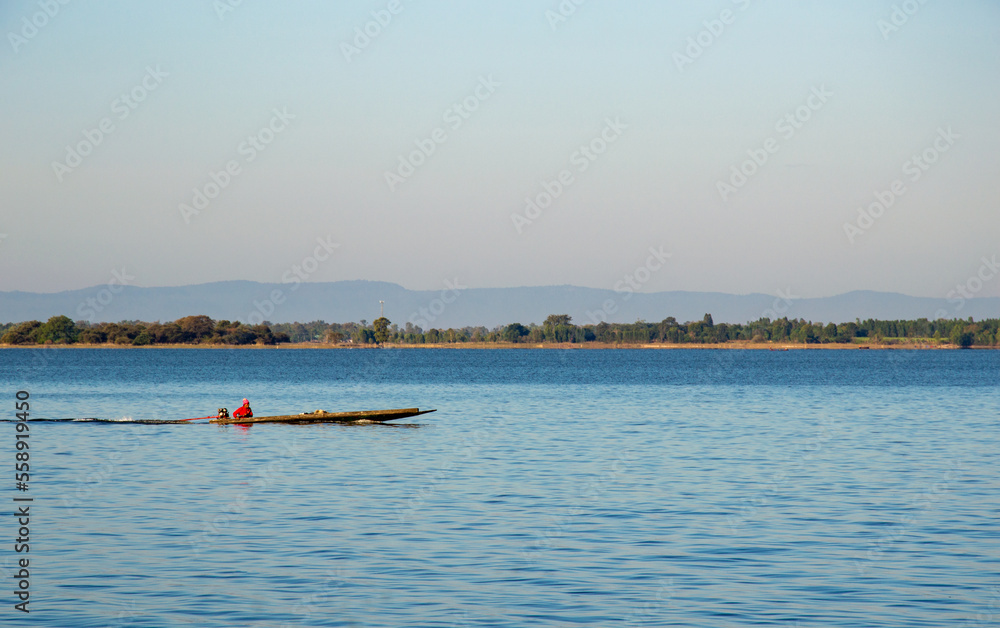 A fisherman wearing a red shirt is driving a boat on the river.  forest and mountain backdrop