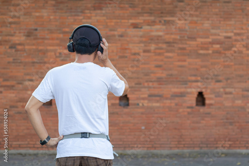 Back view of young man wearing white shirt and black hat holding headphones listening to music via bluetooth happily alone on blurred background , man listening to music with headphones and copy space