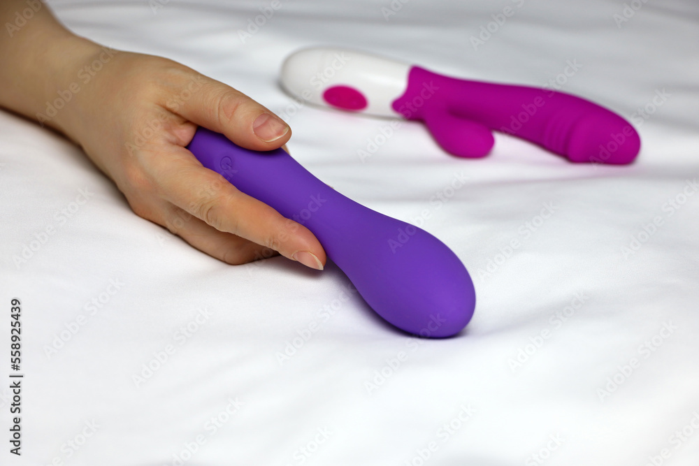 Sex toys in female hand on white sheet. Woman lying on a bed with purple silicone dildo and red vibrator - obrazy, fototapety, plakaty 