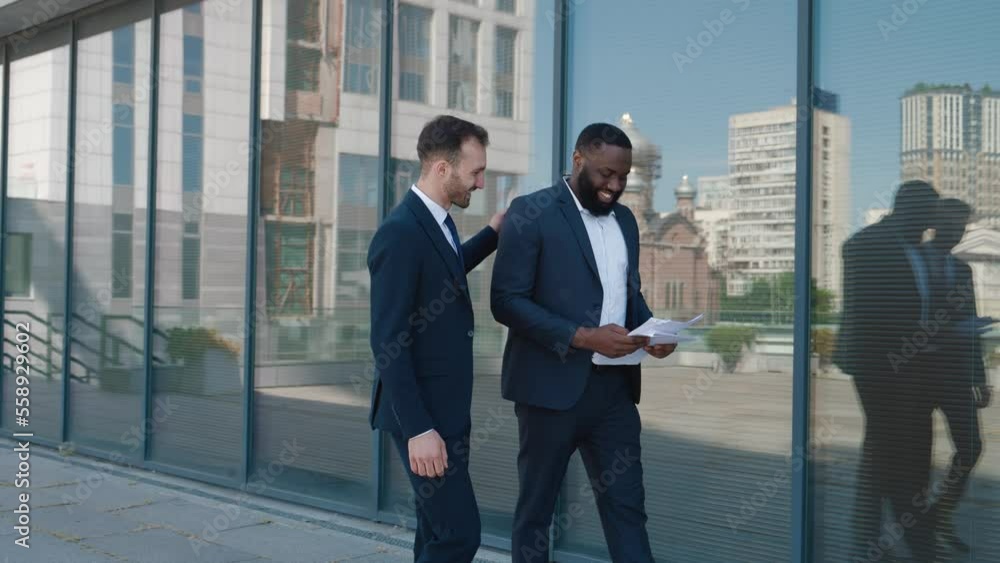 Two smiling males bankers discussing invest collaboration while walking downtown. Two colleagues men bankers in suits talking invest collaboration in financial sector, walking near the office outdoors Two smiling males bankers discussing invest collaboration while walking downtown. Two colleagues men bankers in suits talking invest collaboration in financial sector, walking near the office outdoors