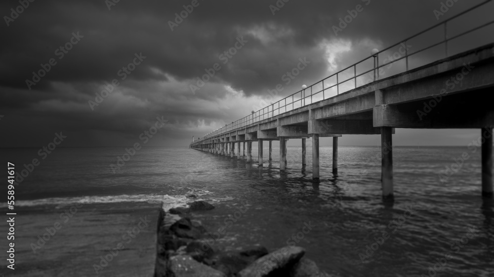 Fototapeta premium Long exposure fine art black and white image of a long pier into the ocean. Silky smooth sea water washes around the concrete pillar. Dark exposure reinforces the gloomy impression. Copy space concept
