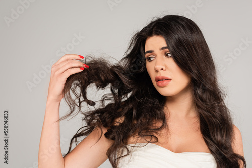 displeased brunette woman looking at long and tousled hair isolated on grey.