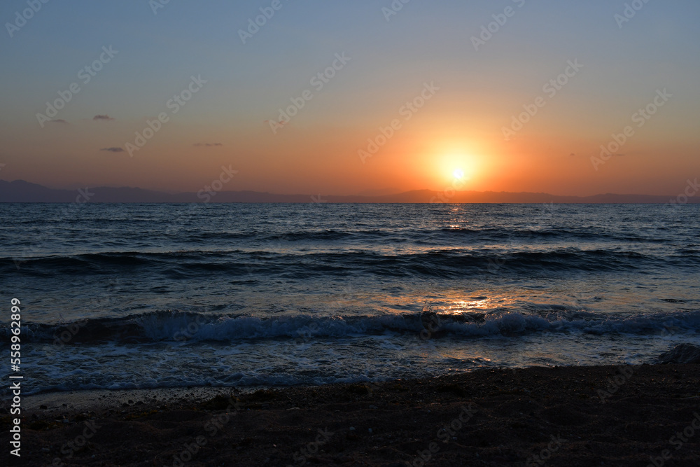 The sun rises over the sea from behind the mountains. the sea surf, small waves rush to the shore. Dawn in dark colors. Reflection of the sun on the water surface. Beach at dawn