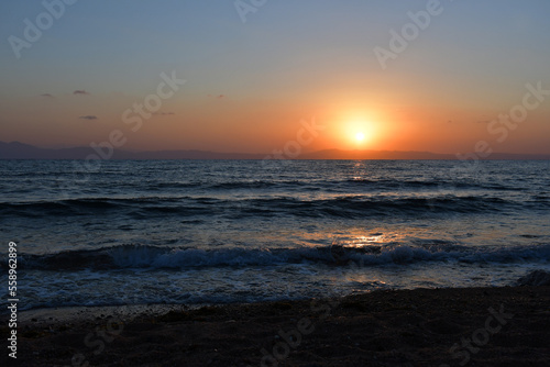 The sun rises over the sea from behind the mountains. the sea surf, small waves rush to the shore. Dawn in dark colors. Reflection of the sun on the water surface. Beach at dawn