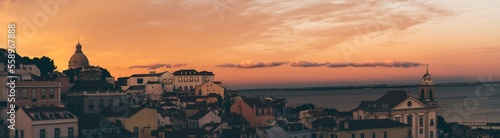 A panoramic image with a cityscape of an antique part of Lisbon with old houses, churches, and a river with a stunning sunset in the background; a panorama of an old Portuguese city, dramatic evening