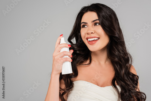 joyful woman with shiny hair holding treatment spray bottle isolated on grey.