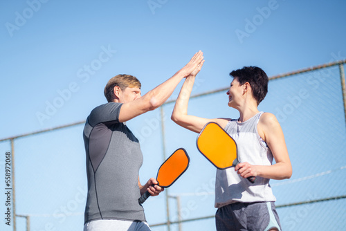 Laughing couple playing pickleball game, hitting pickleball yellow ball with paddle, outdoor sport leisure activity, celebrating victory .