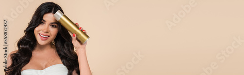 Smiling brunette woman holding hairspray isolated on beige with copy space, banner.