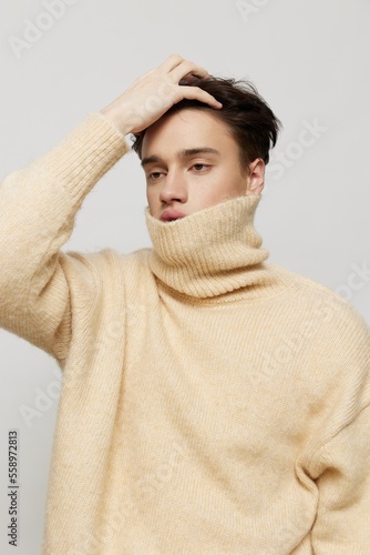 A handsome young man with dark short hair combed back in a beige sweater with a high neck stands on a gray background with a hand on his head .Vertical studio shot. © Tatiana