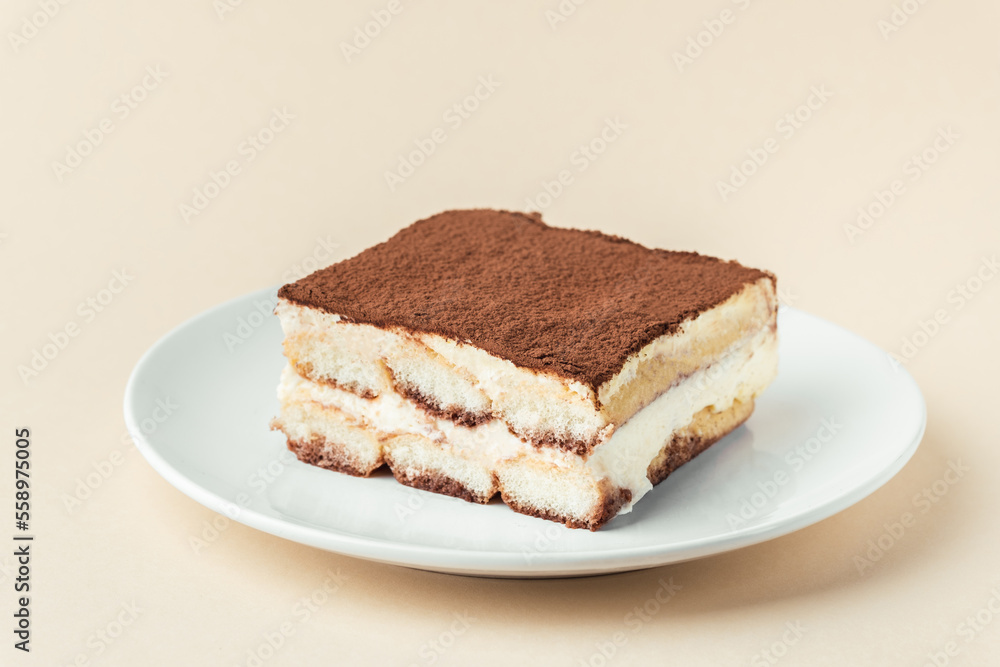 Homemade tiramisu cake on white plate against beige background. Traditional Italian no-bake dessert made of savoiardi, filled with mascarpone cheese, coffee espresso and sprinkled with cocoa powder Homemade tiramisu cake on white plate against beige background. Traditional Italian no-bake dessert made of savoiardi, filled with mascarpone cheese, coffee espresso and sprinkled with cocoa powder