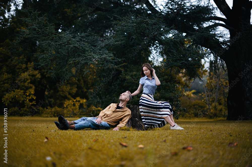 A romantic photo of a young couple in a beautiful autumn park, with the woman sitting on a tree stump and the man lying nearby A romantic photo of a young couple in a beautiful autumn park, with the woman sitting on a tree stump and the man lying nearby